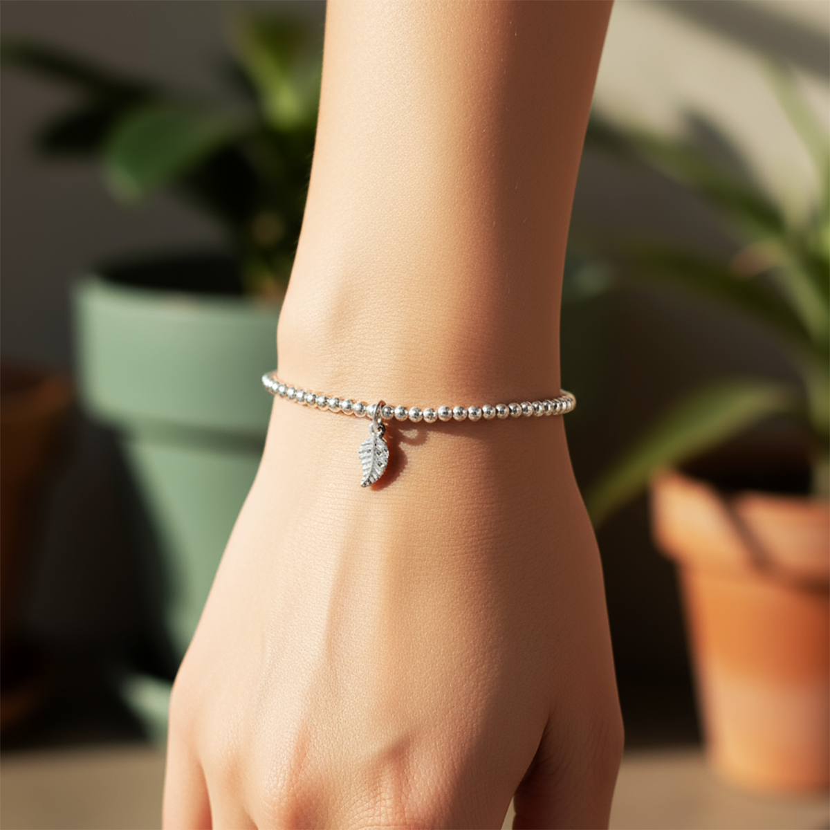 Close-up of a hand wearing a silver bracelet with a leaf charm, blurred plants in the background