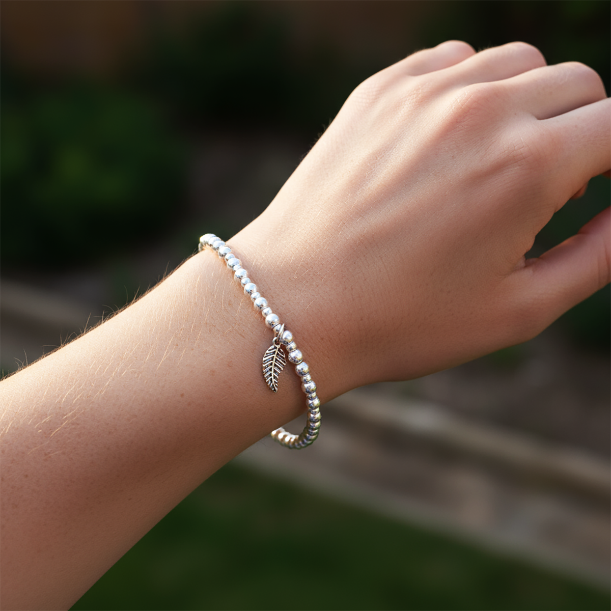 Hand wearing a beaded bracelet with a leaf charm against a blurred natural background