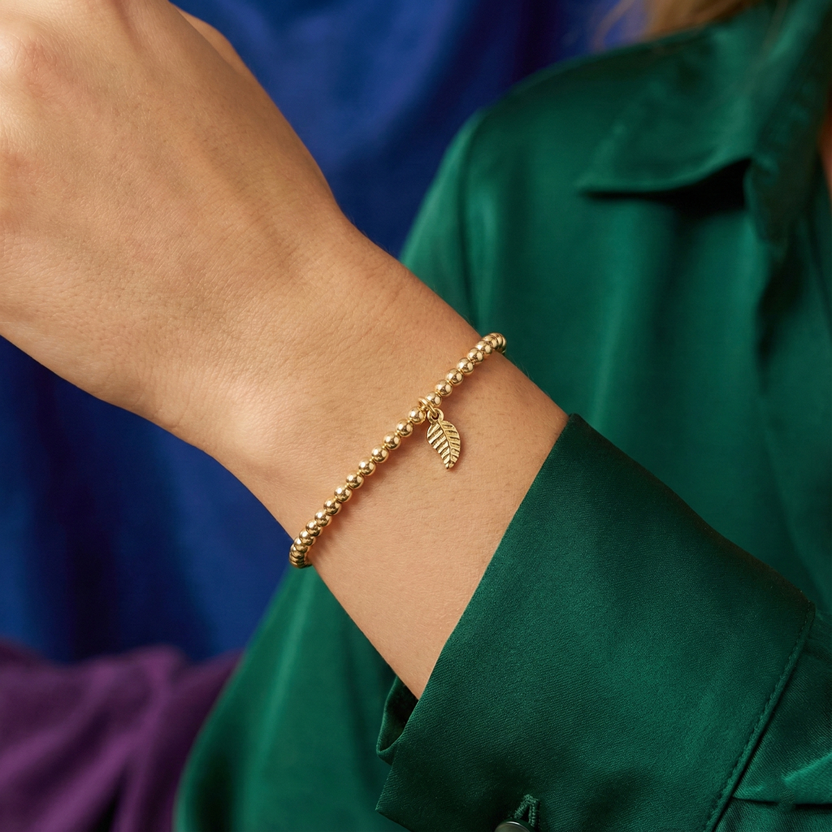 Gold bracelet with leaf charm on a wrist against a green and blue background