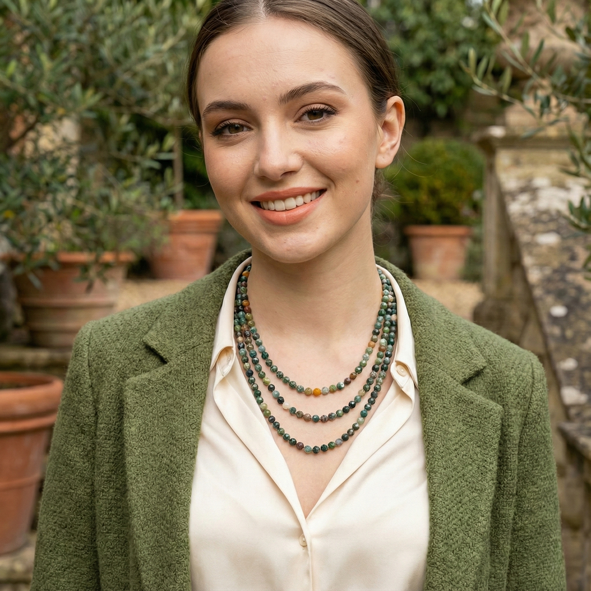 Woman wearing a green blazer and multiple necklaces outdoors with plants in the background