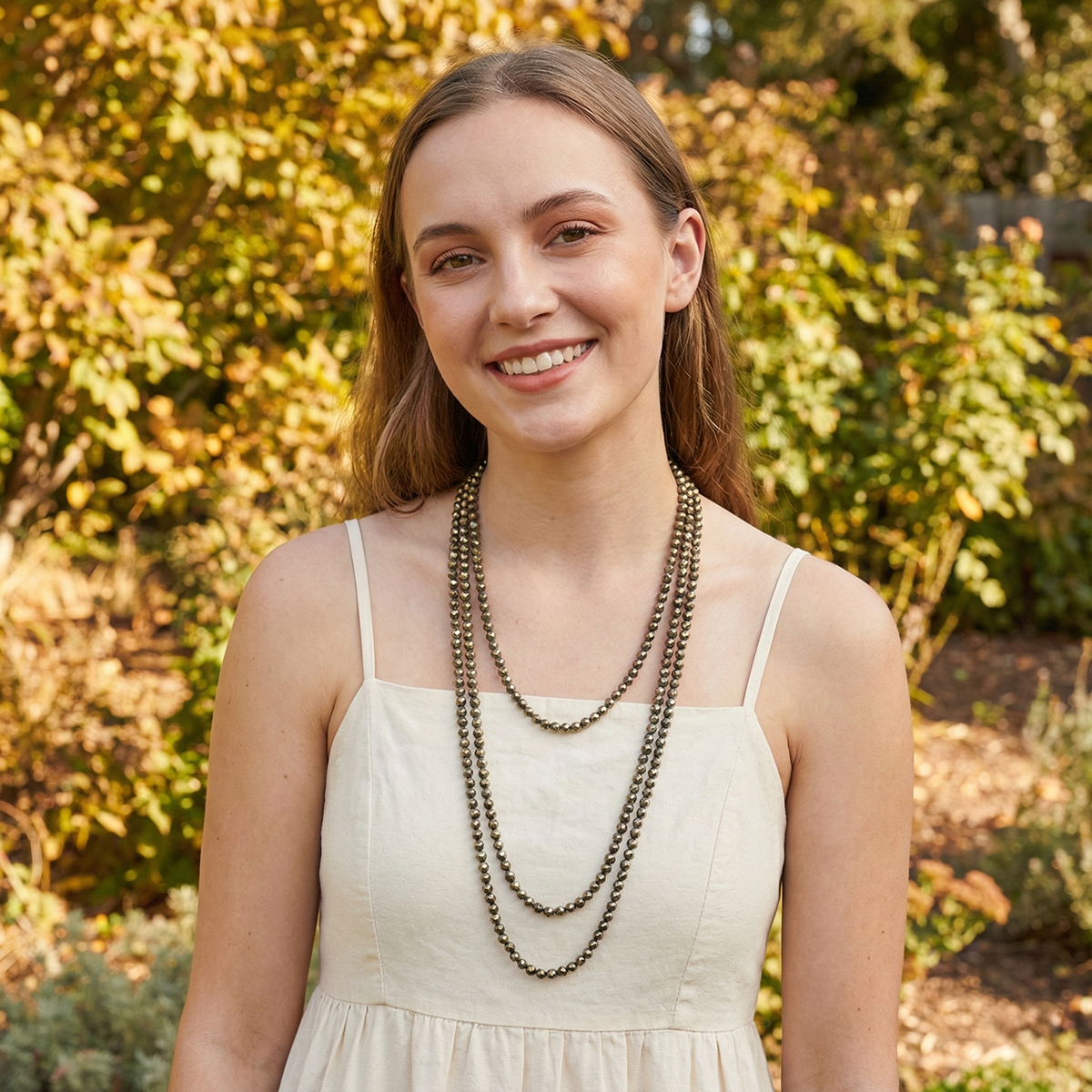 Woman wearing a white dress and multiple necklaces standing outdoors with greenery in the background