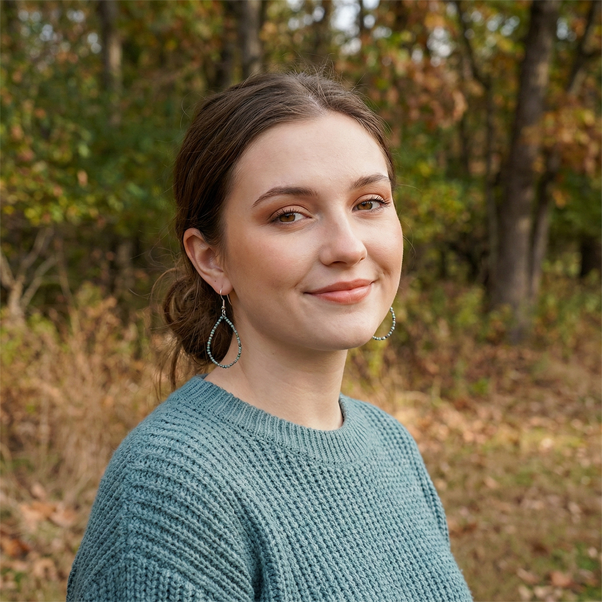 Woman in a teal sweater standing in an outdoor setting with trees and foliage.
