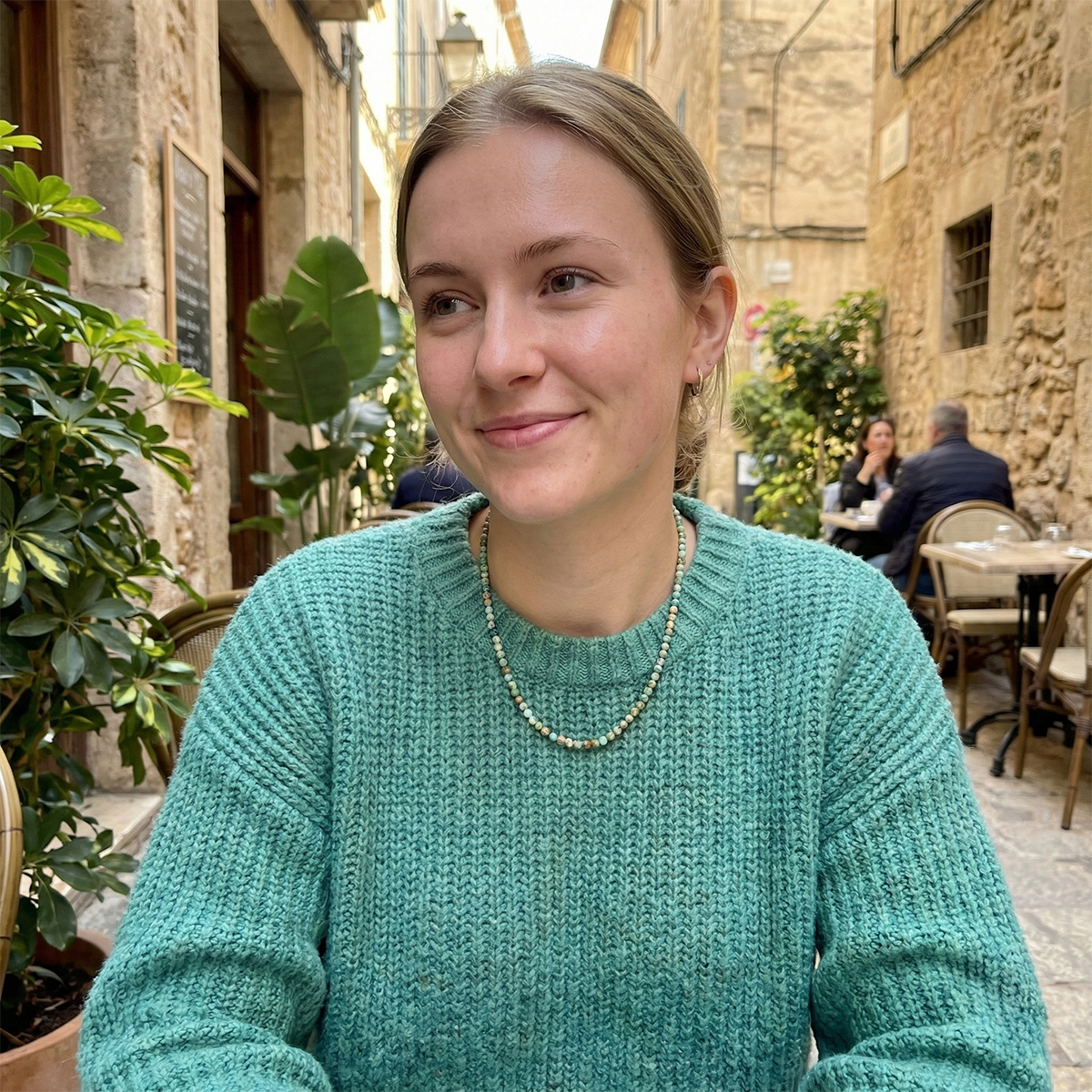 Woman in a teal sweater sitting outdoors with stone buildings and plants in the background