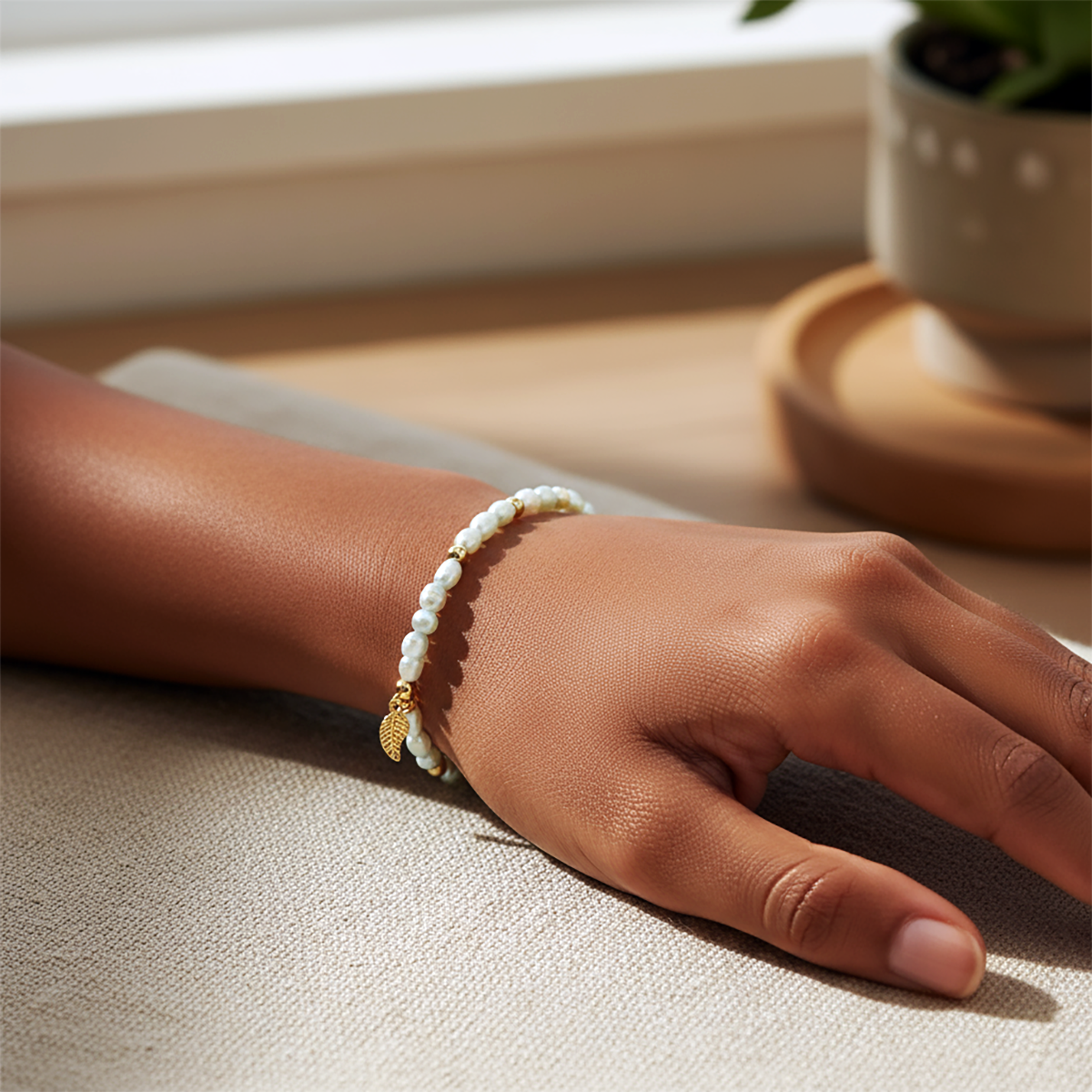 Hand wearing a pearl bracelet on a light surface with a plant in the background