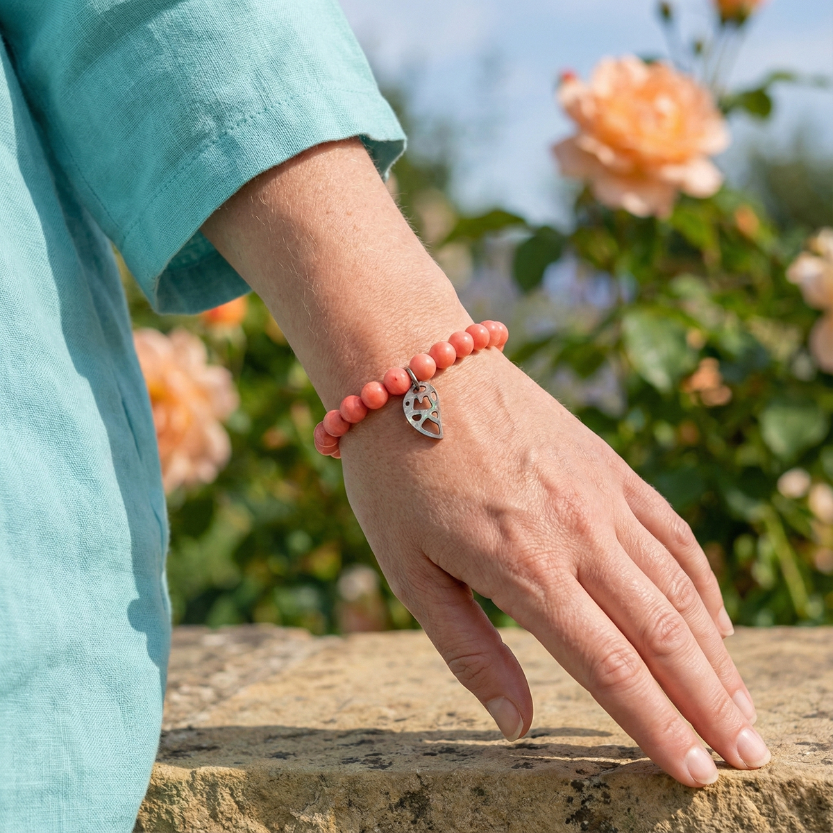 Hand wearing a red beaded bracelet with a silver heart charm, against a floral background.