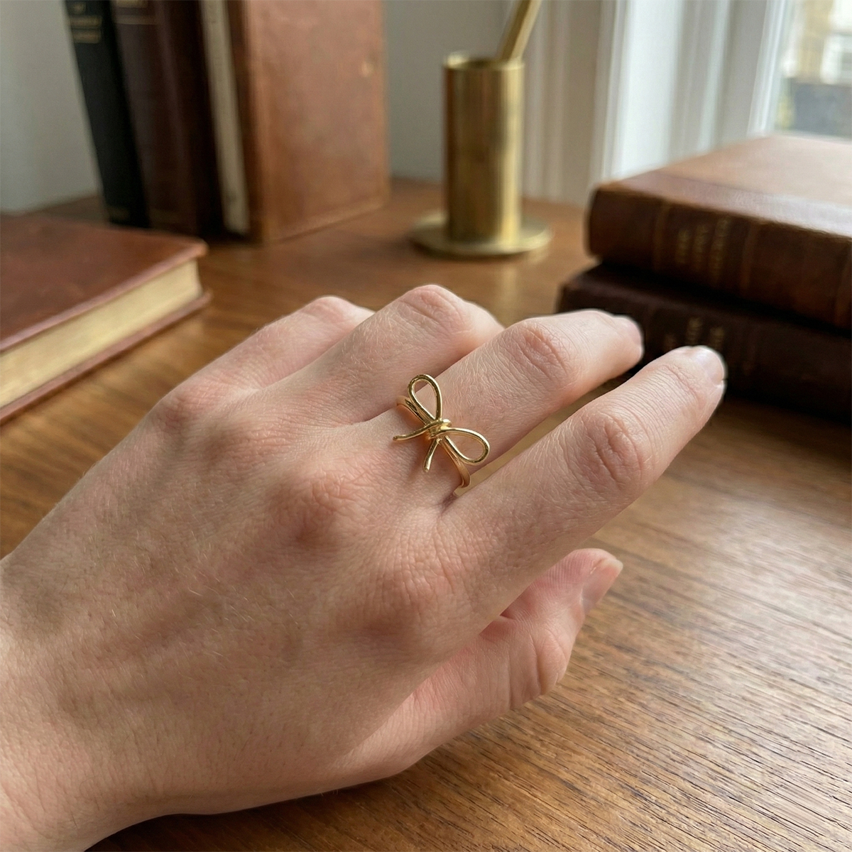 Hand wearing a gold bow-shaped ring on a wooden surface with books and a candle in the background