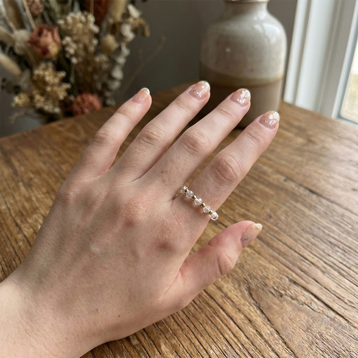 Hand wearing a pearl ring on a wooden surface with a vase and flowers in the background