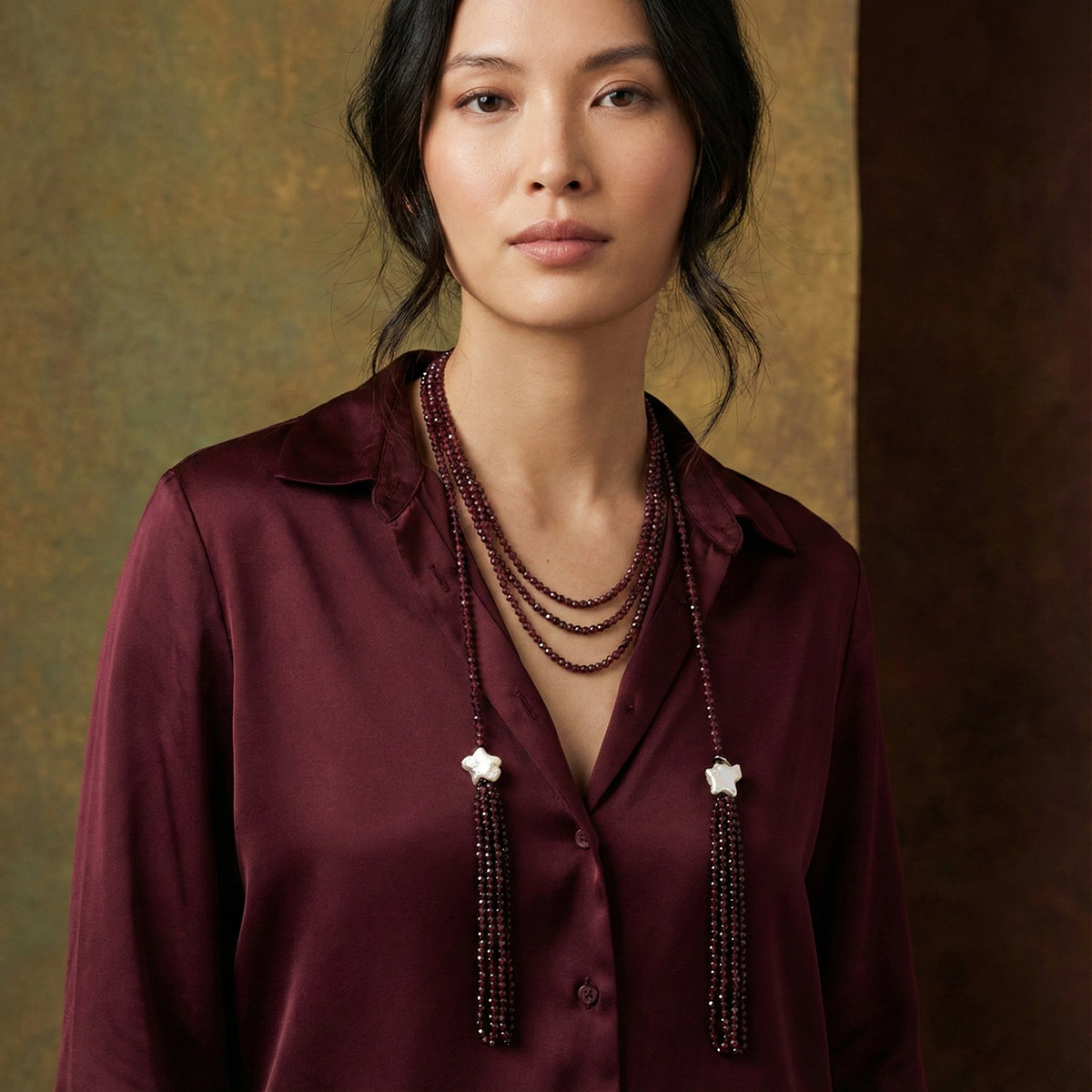 Woman wearing a burgundy shirt and long beaded necklace against a textured wall.