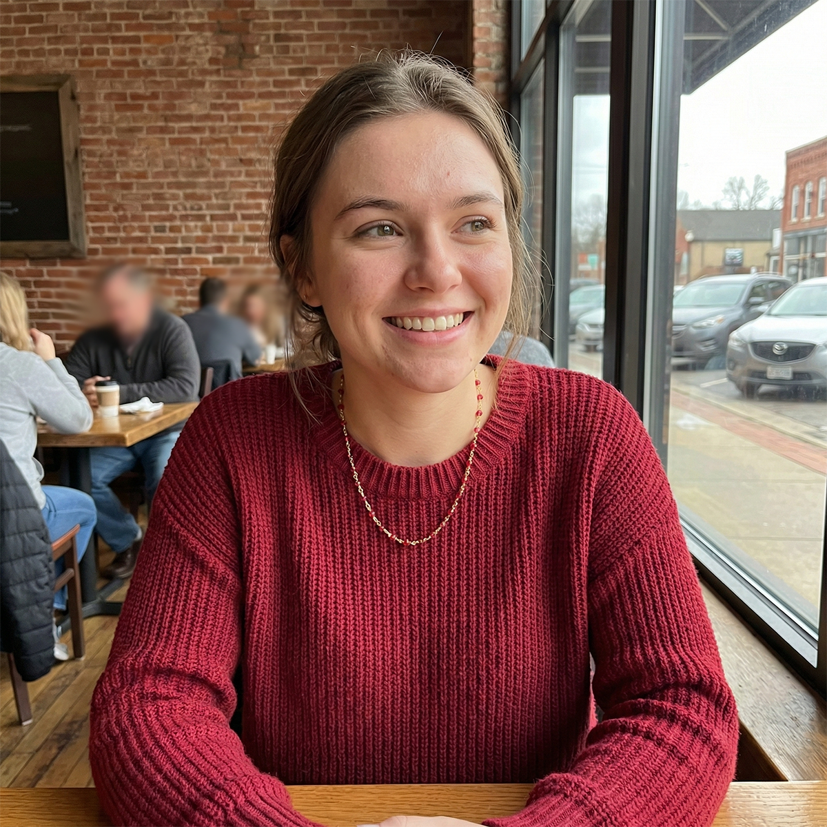 Woman in a red sweater sitting at a table in a casual setting with a brick wall and window in the background.