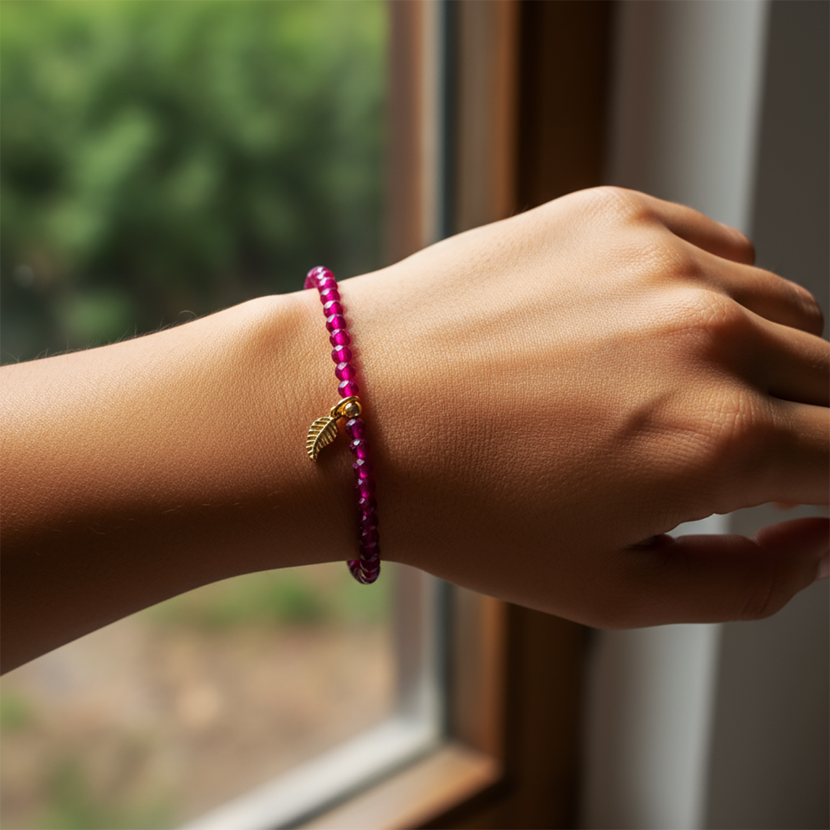 Hand wearing a pink beaded bracelet with a gold charm by a window