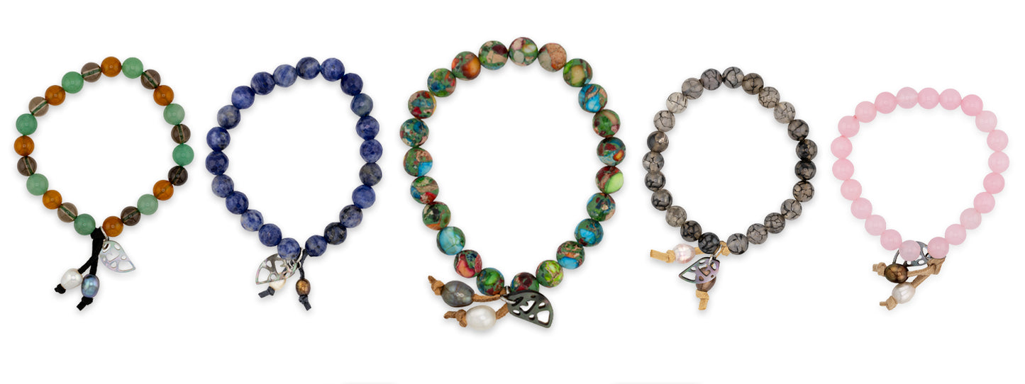 Five beaded bracelets arranged in a row on a white background, each made of round gemstone-like beads and featuring small hanging charms. The bracelets include green and orange, dark blue, multicolored iridescent, gray, and light pink beads.