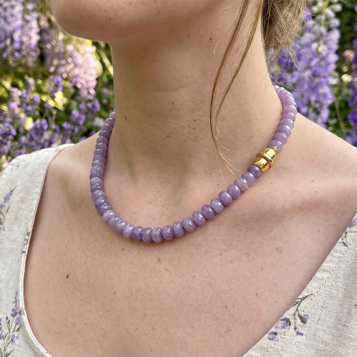 Person wearing a purple beaded necklace with a gold clasp against a blurred floral background