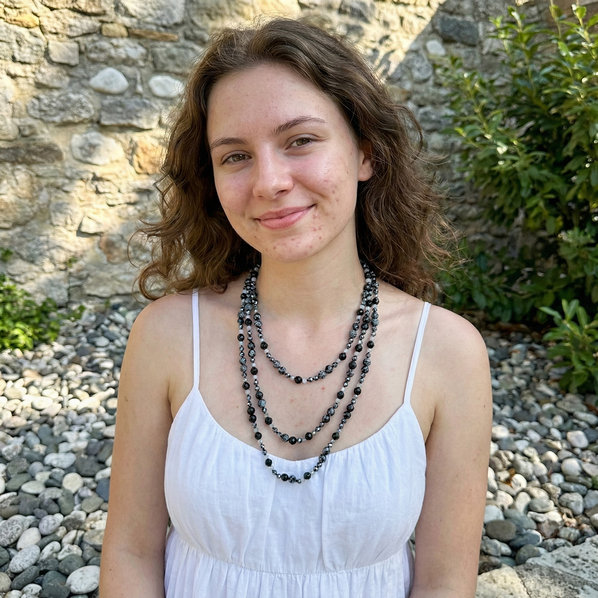Woman wearing a white dress and multiple necklaces against a stone wall background