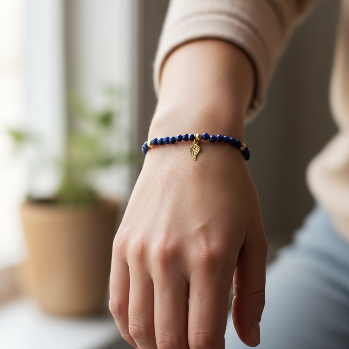 Hand wearing a blue beaded bracelet with a leaf charm, blurred indoor background