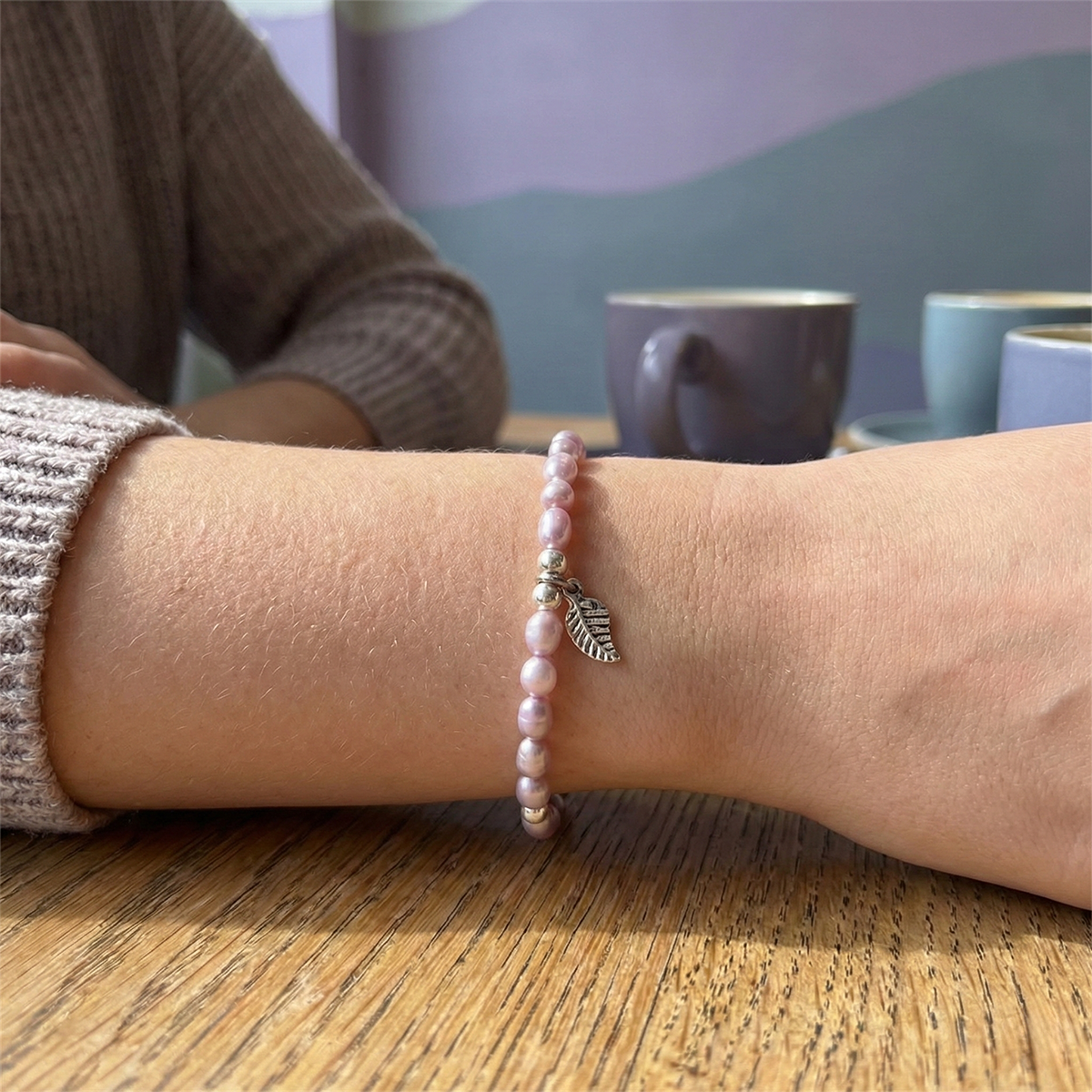 Person wearing a pink beaded bracelet with a leaf charm on a wooden table.