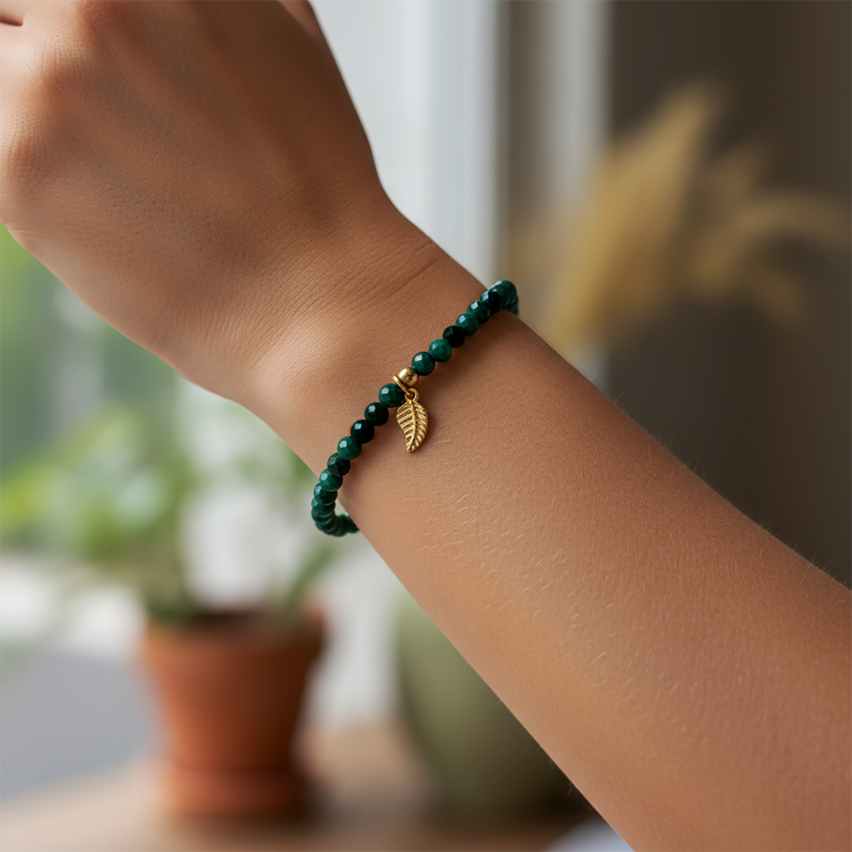 Green beaded bracelet with a gold leaf charm on a wrist, blurred indoor background