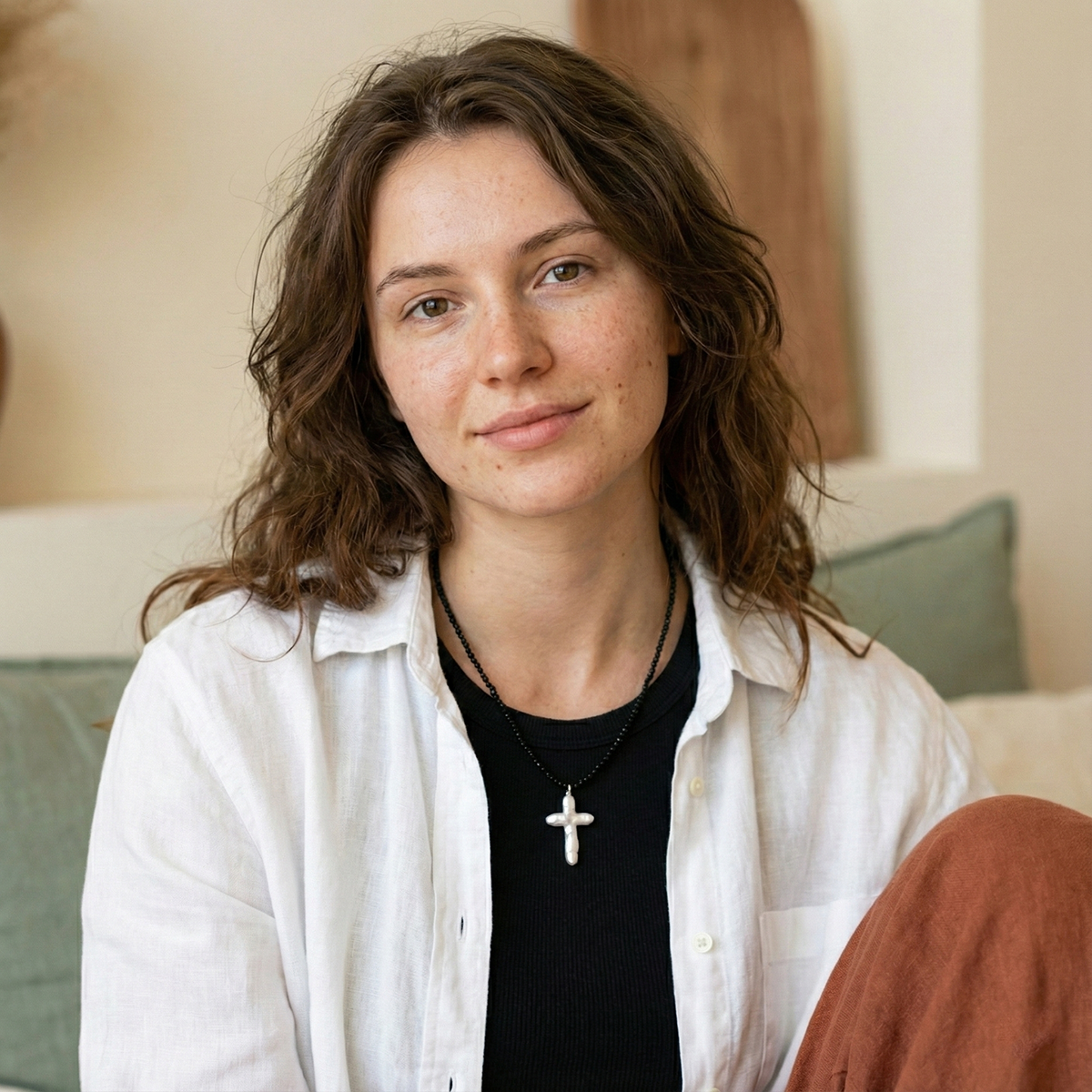 Woman wearing a white shirt and black top with a cross necklace, sitting on a couch.