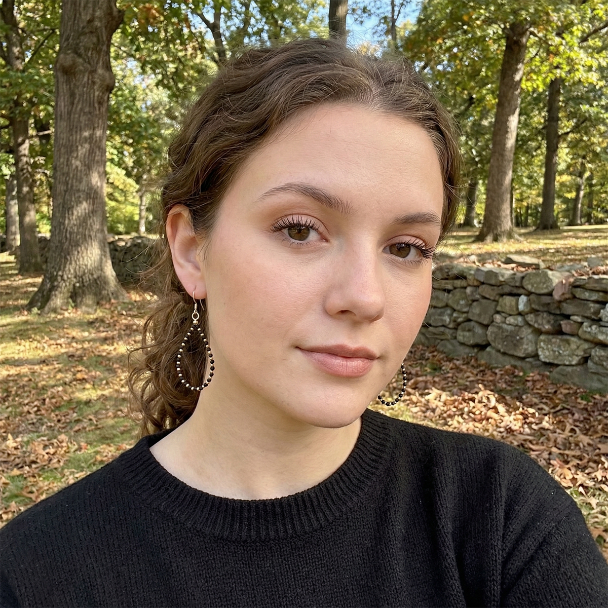 Woman with earrings standing outdoors in a park with trees and a stone wall in the background