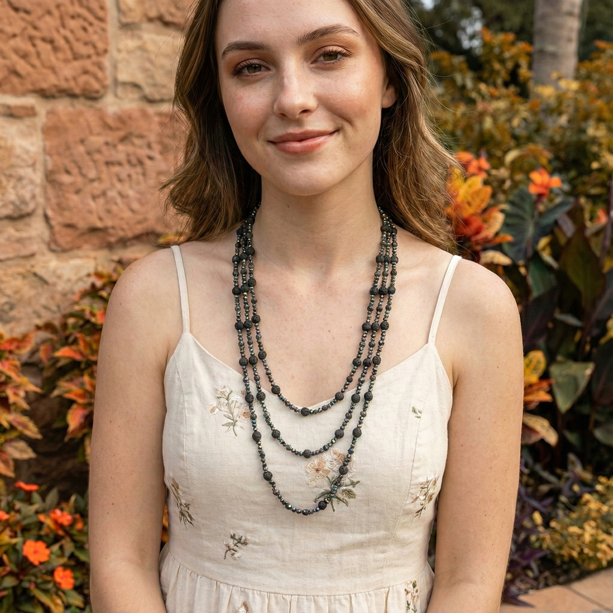 Woman wearing a long black beaded necklace in front of a stone wall and plants.