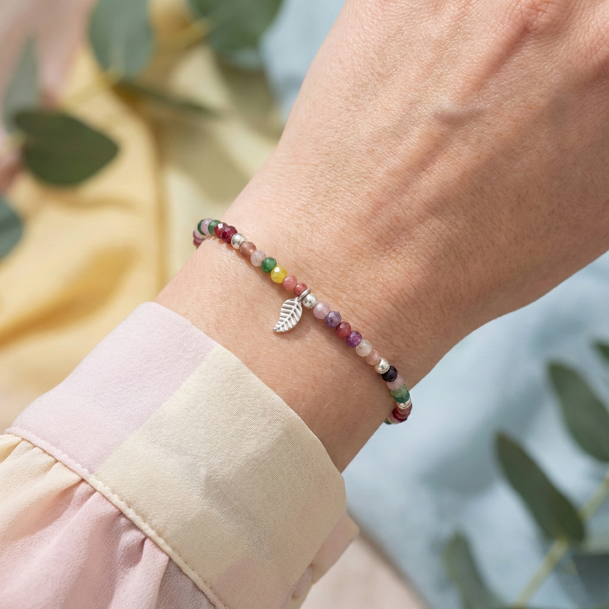 Multicolored beaded bracelet with a leaf charm on a wrist, blurred greenery in the background