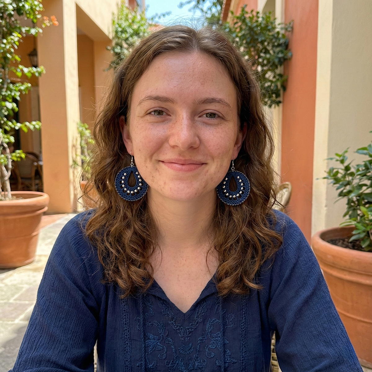 Woman wearing blue earrings and a blue top outdoors with plants in the background