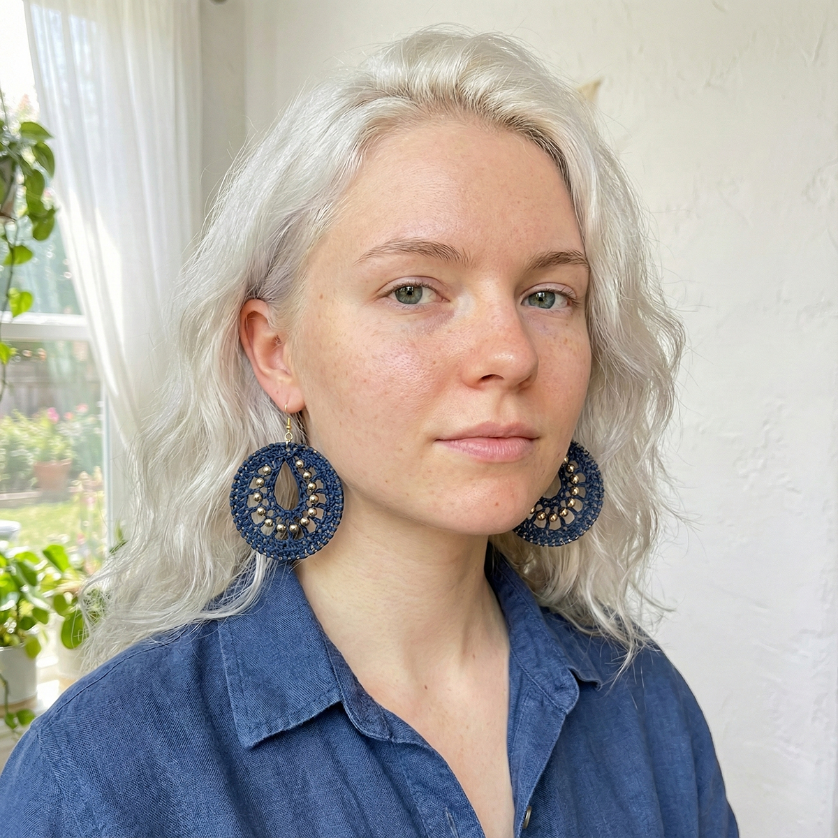 Woman wearing large blue earrings indoors with a window and plants in the background