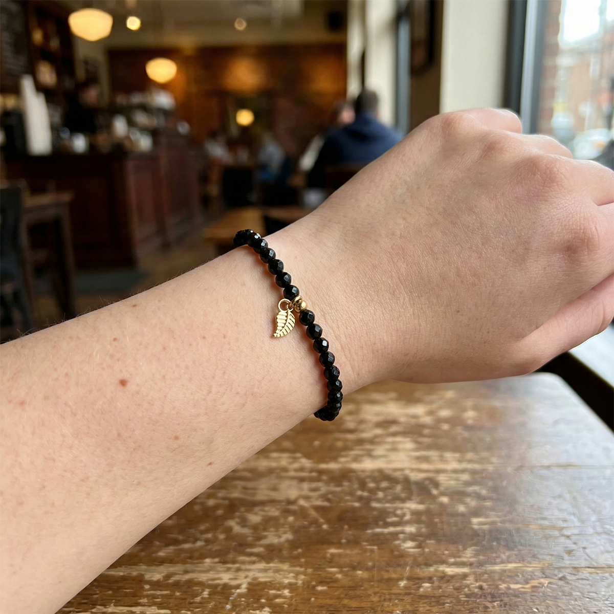 Person wearing a black beaded bracelet with a gold charm on a wooden table in a blurred cafe setting