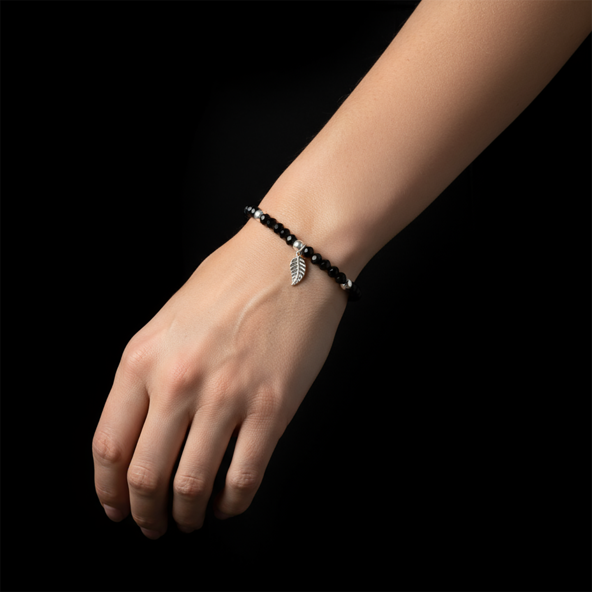 Hand wearing a black beaded bracelet with a leaf charm on a black background