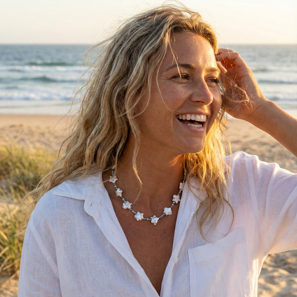 Woman on a beach wearing a white shirt and a necklace with starfish, smiling.