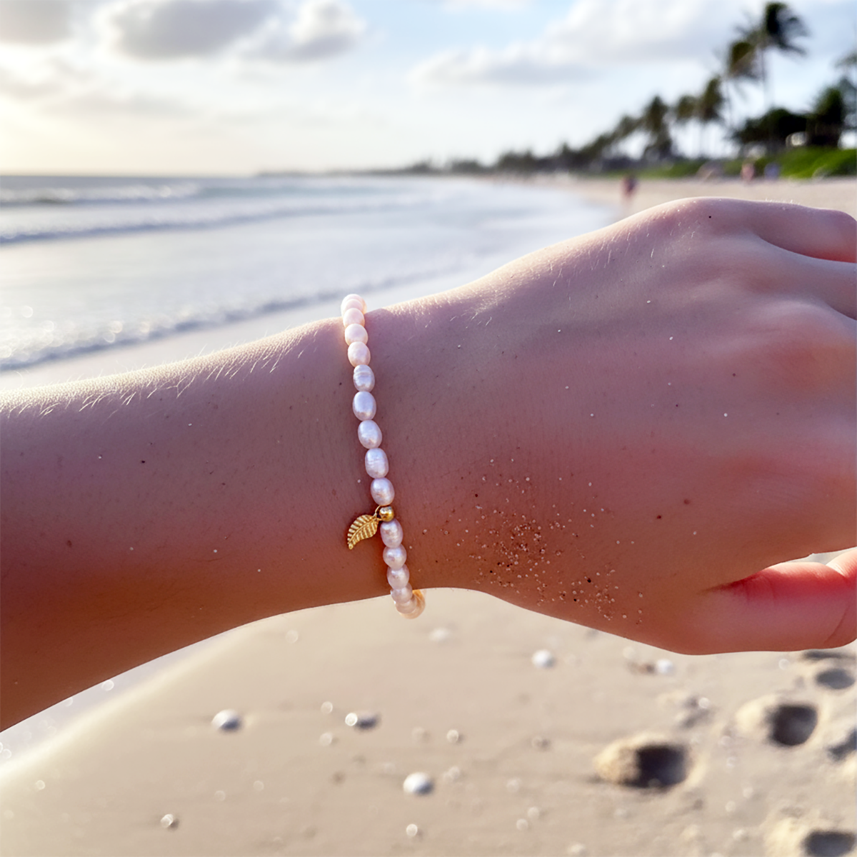 Hand wearing a pearl bracelet on a beach with palm trees in the background