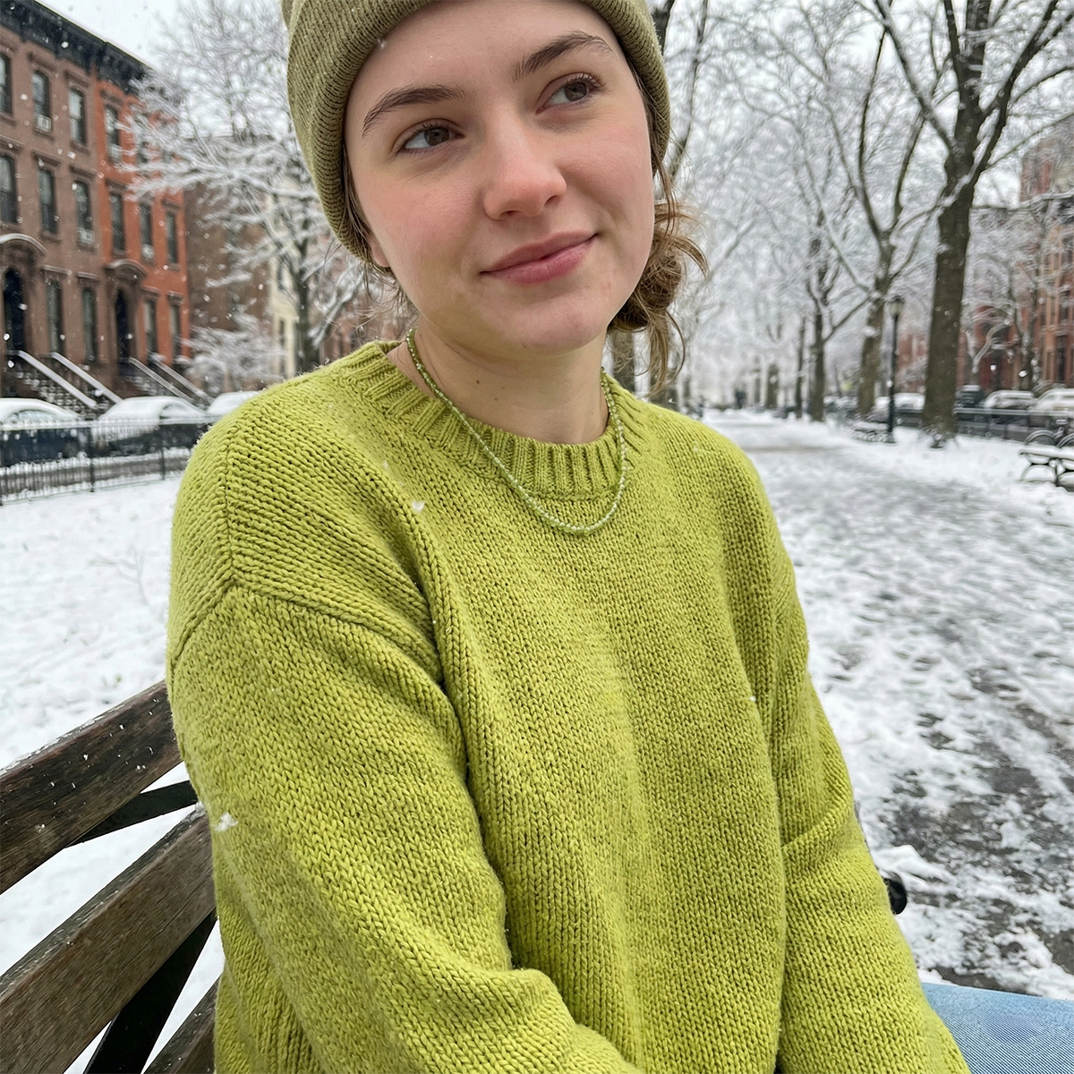 Person wearing a green sweater and beanie sitting on a snowy bench.