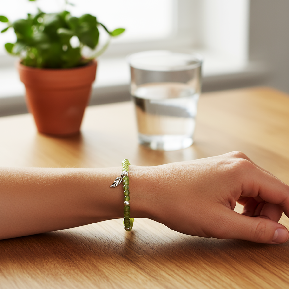 Hand wearing a green bracelet on a wooden table with a plant and glass in the background