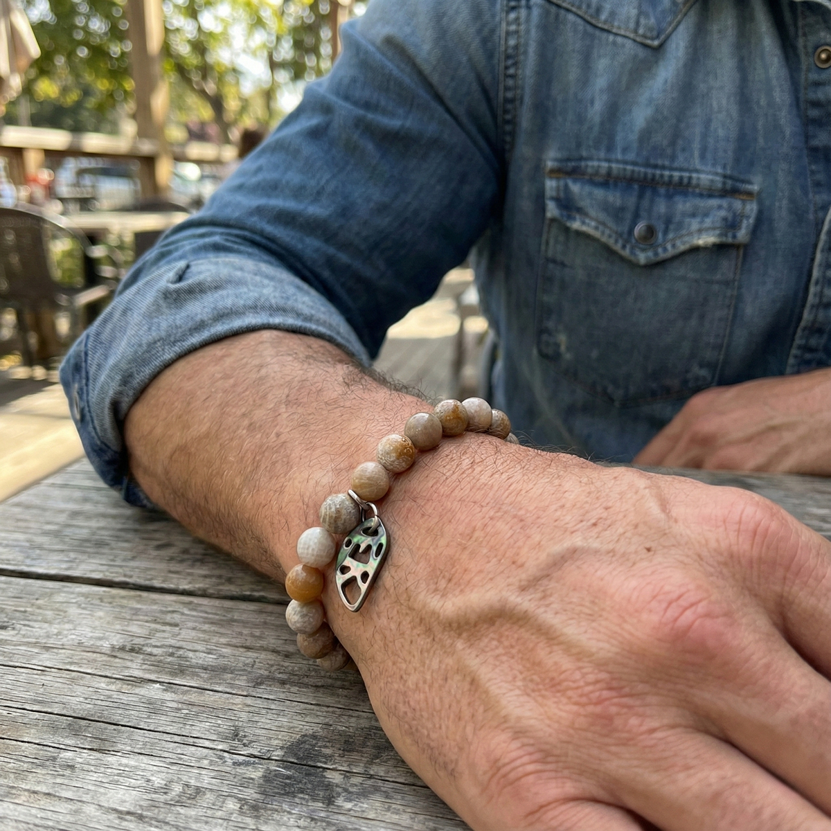 Person wearing a beaded bracelet with a heart charm on a wooden table outdoors.