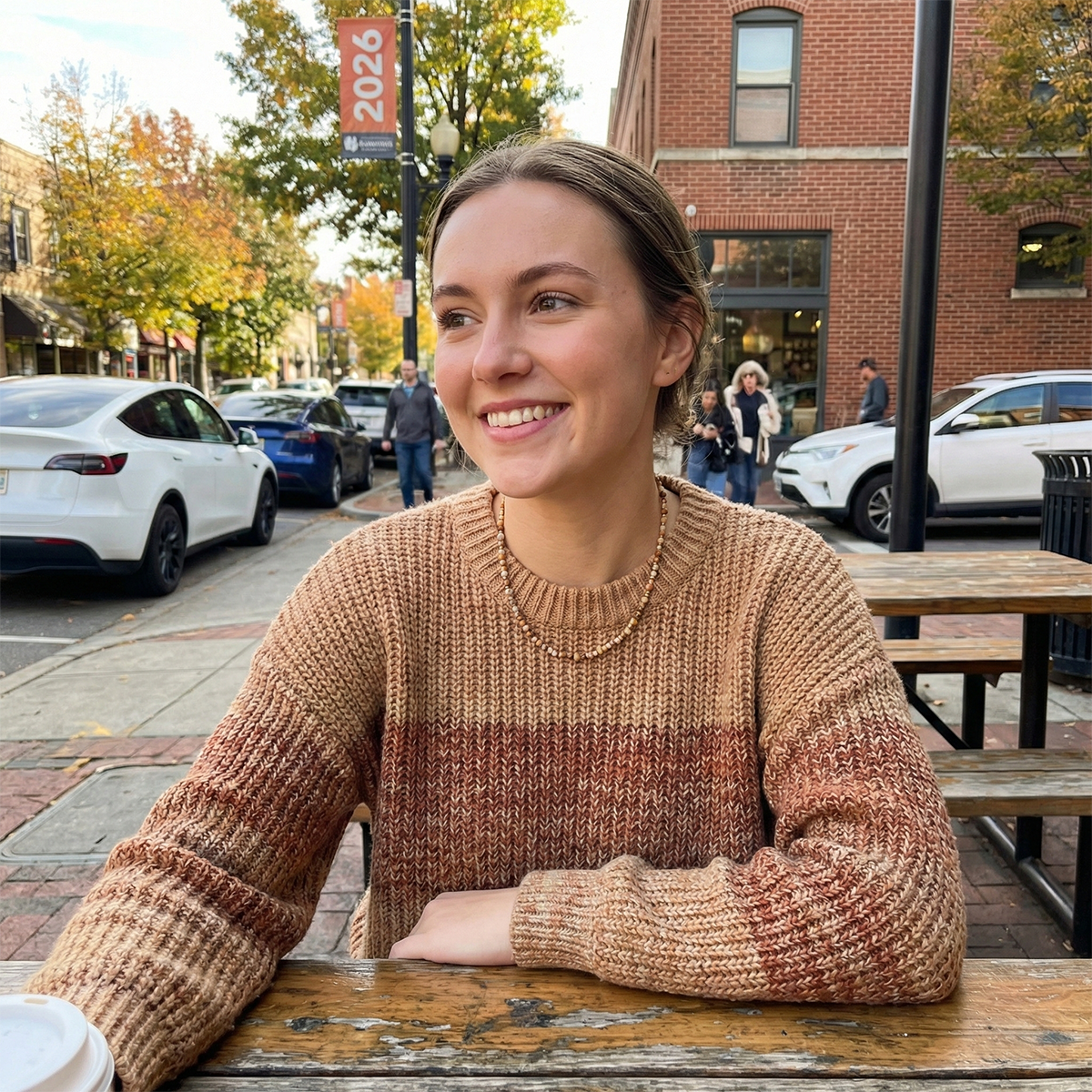 Woman sitting at an outdoor table in a city street, wearing a brown sweater.