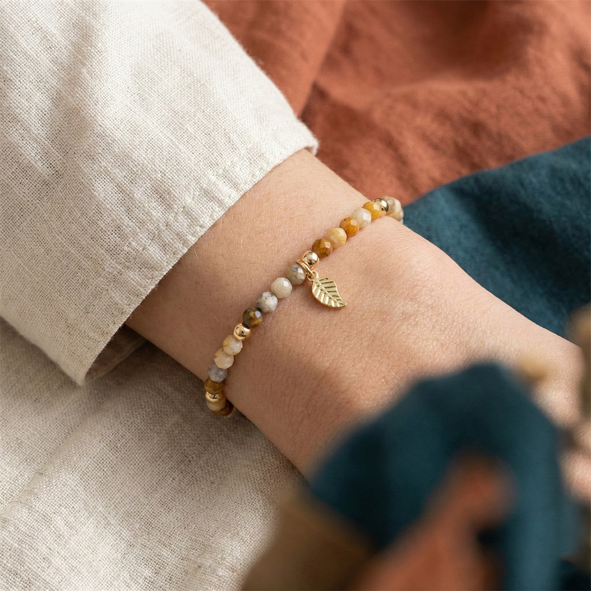 Close-up of a wrist wearing a beaded bracelet with a gold leaf charm on a textured fabric background.