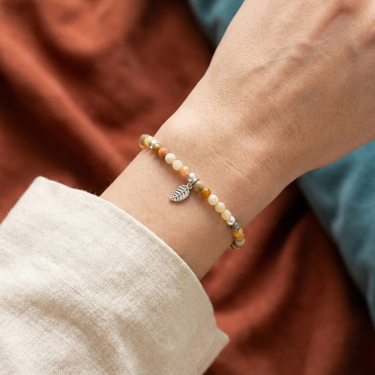Close-up of a wrist wearing a beaded bracelet with a leaf charm against a warm-toned fabric background.