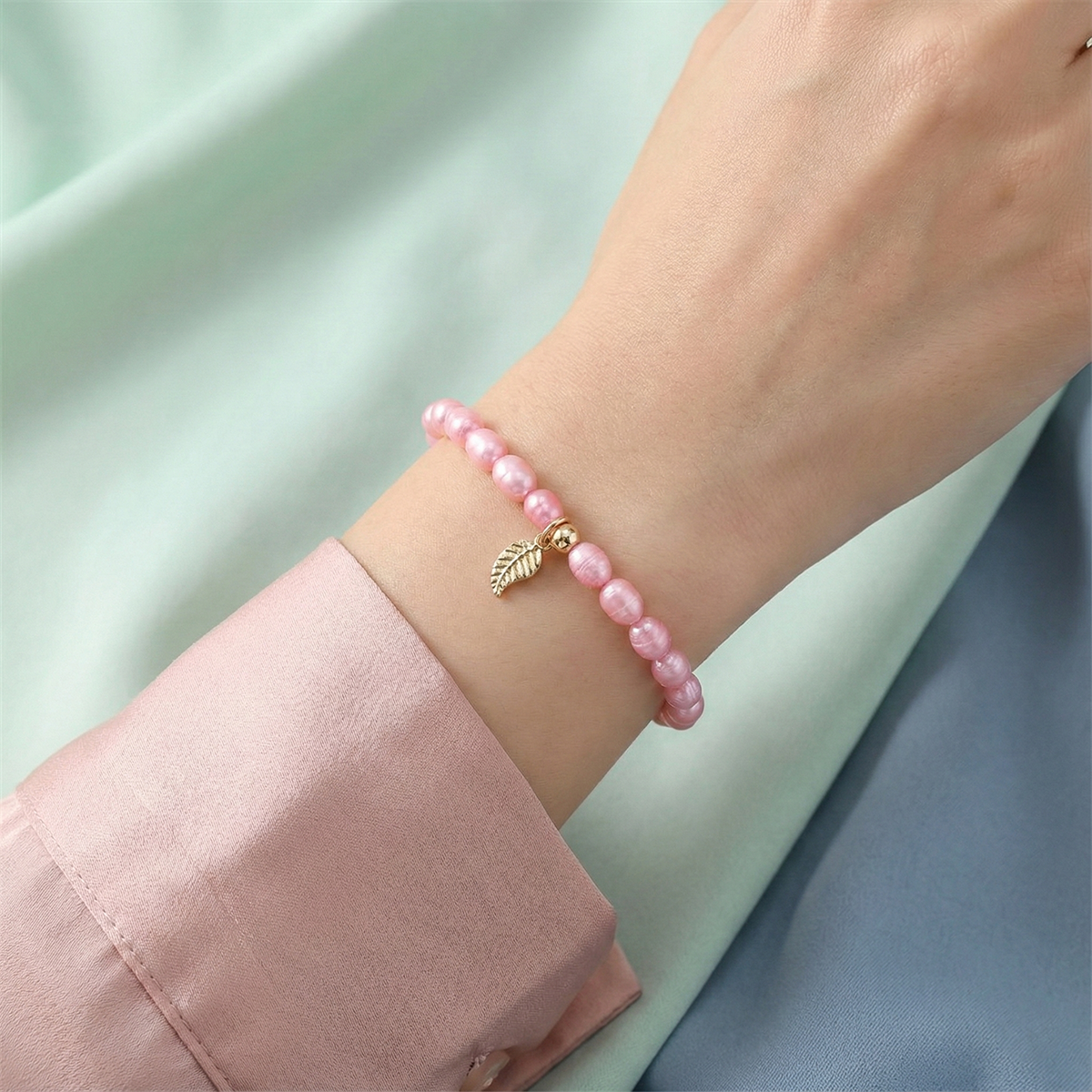 Pink beaded bracelet with a gold leaf charm on a wrist against a light green background