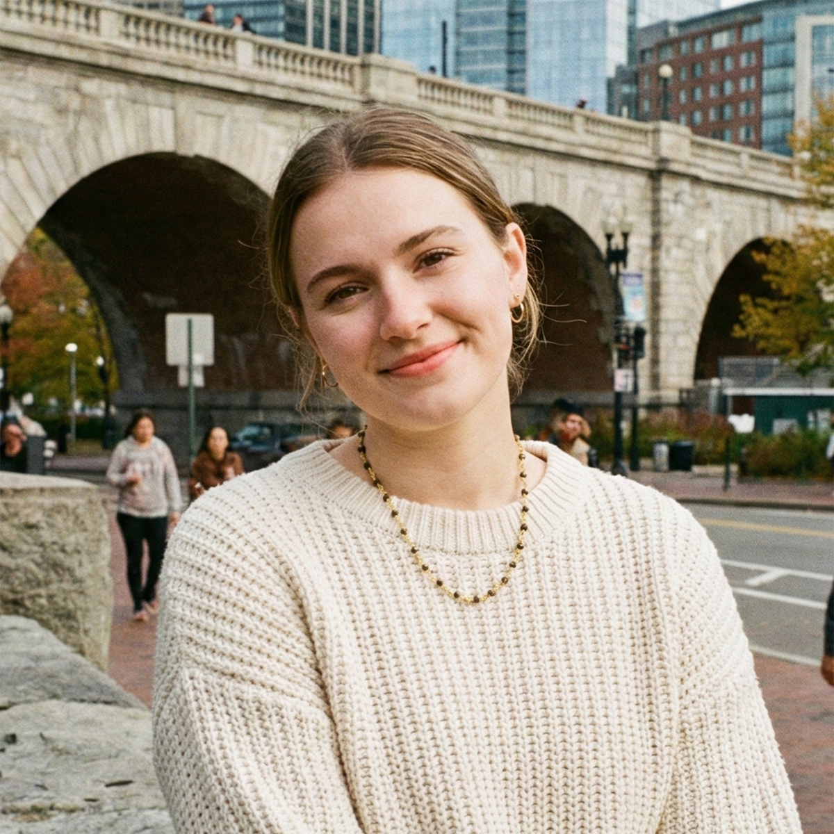 Woman in a beige sweater standing in front of a stone arch bridge with people and buildings in the background.