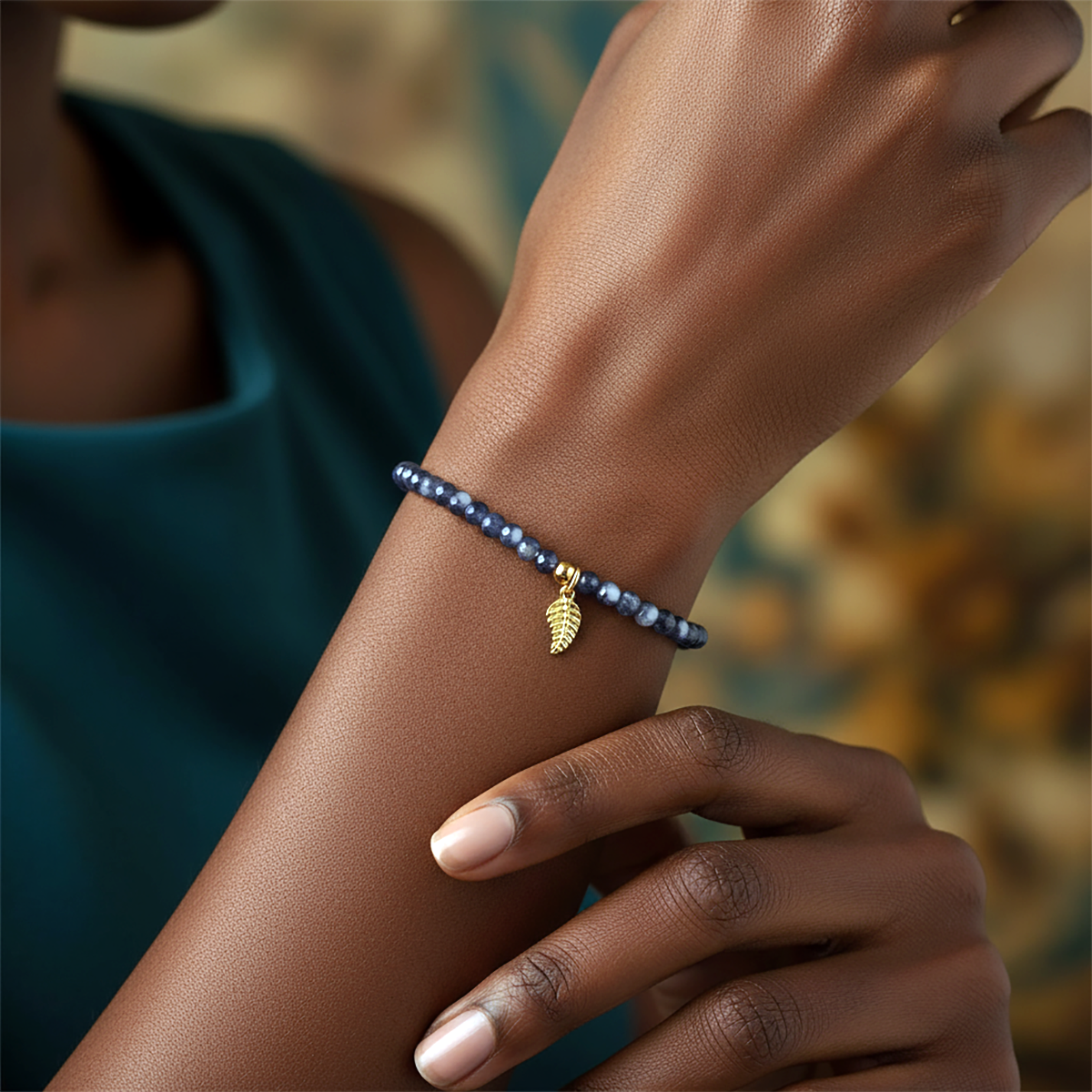 Close-up of a hand wearing a blue beaded bracelet with a gold leaf charm against a blurred background.
