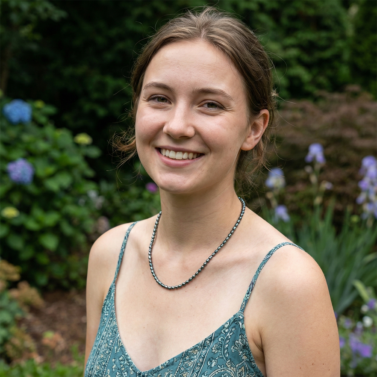 Woman wearing a green patterned dress with a necklace, standing in a garden with flowers and plants.