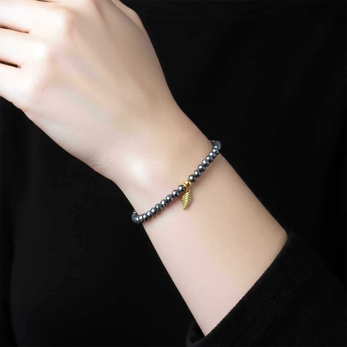 Hand wearing a bracelet with a gold leaf charm on a black background