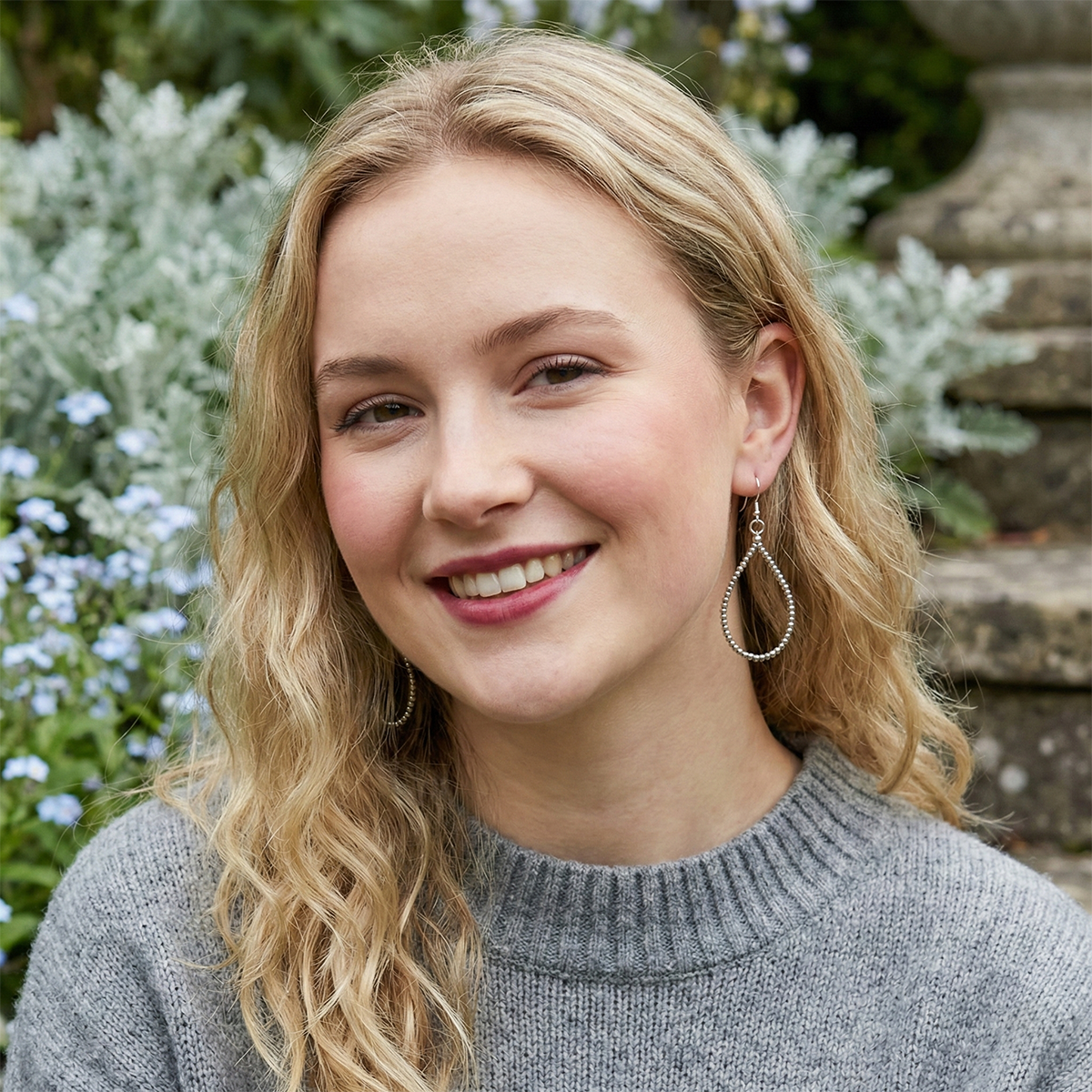 Woman with blonde hair and earrings smiling outdoors with greenery in the background