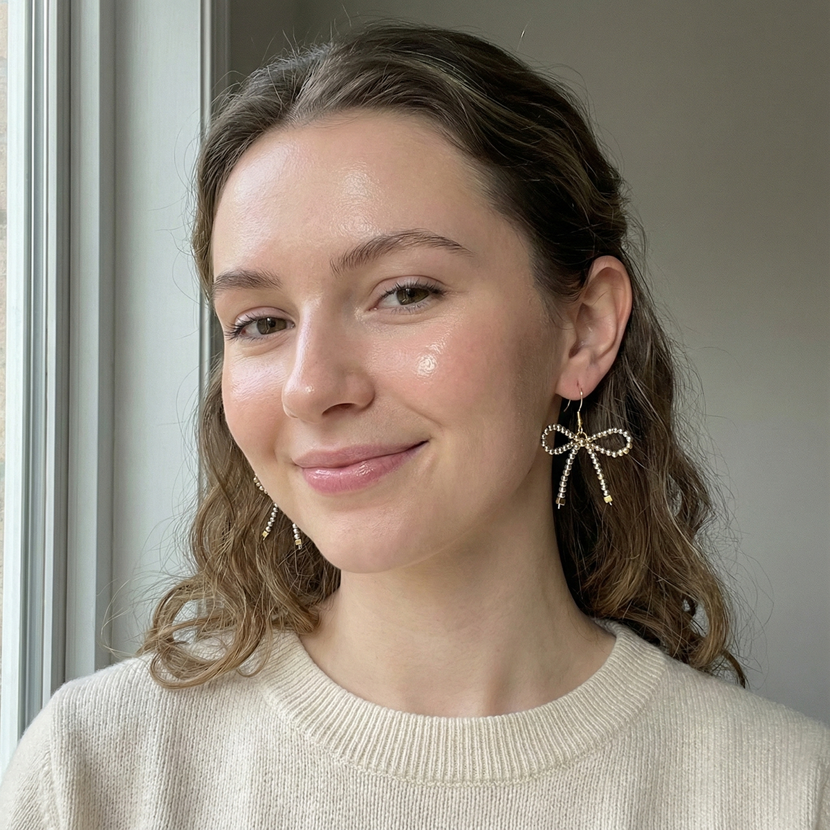 Woman wearing a beige sweater and bow-shaped earrings indoors.