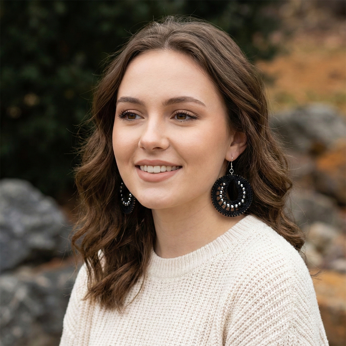 Woman with long brown hair wearing a beige sweater and black earrings outdoors.