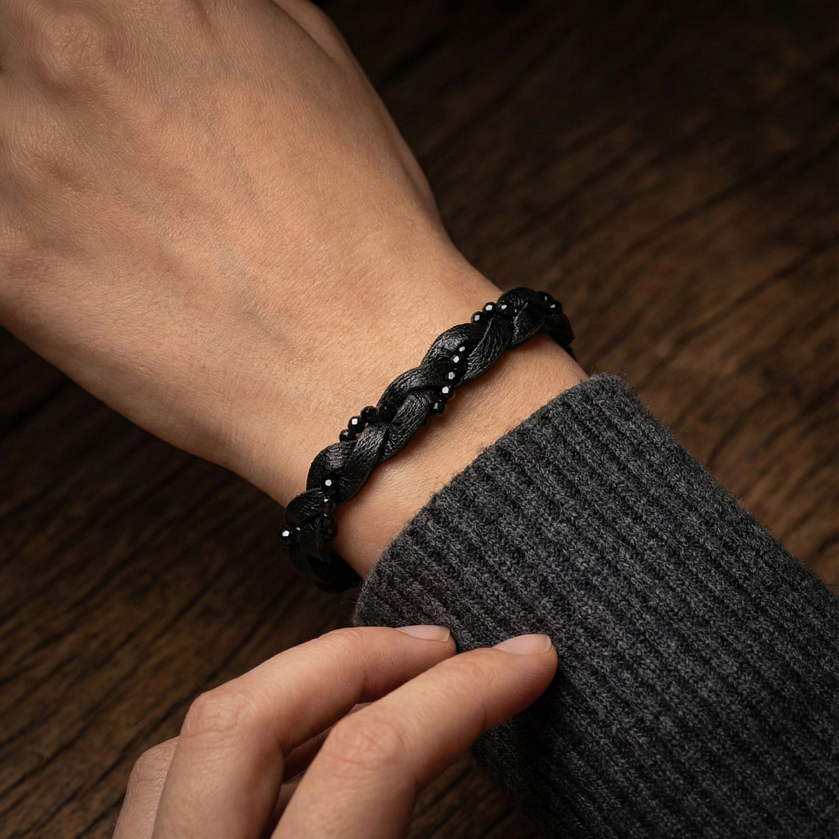 Black braided bracelet on a wrist with a dark sweater sleeve against a wooden background