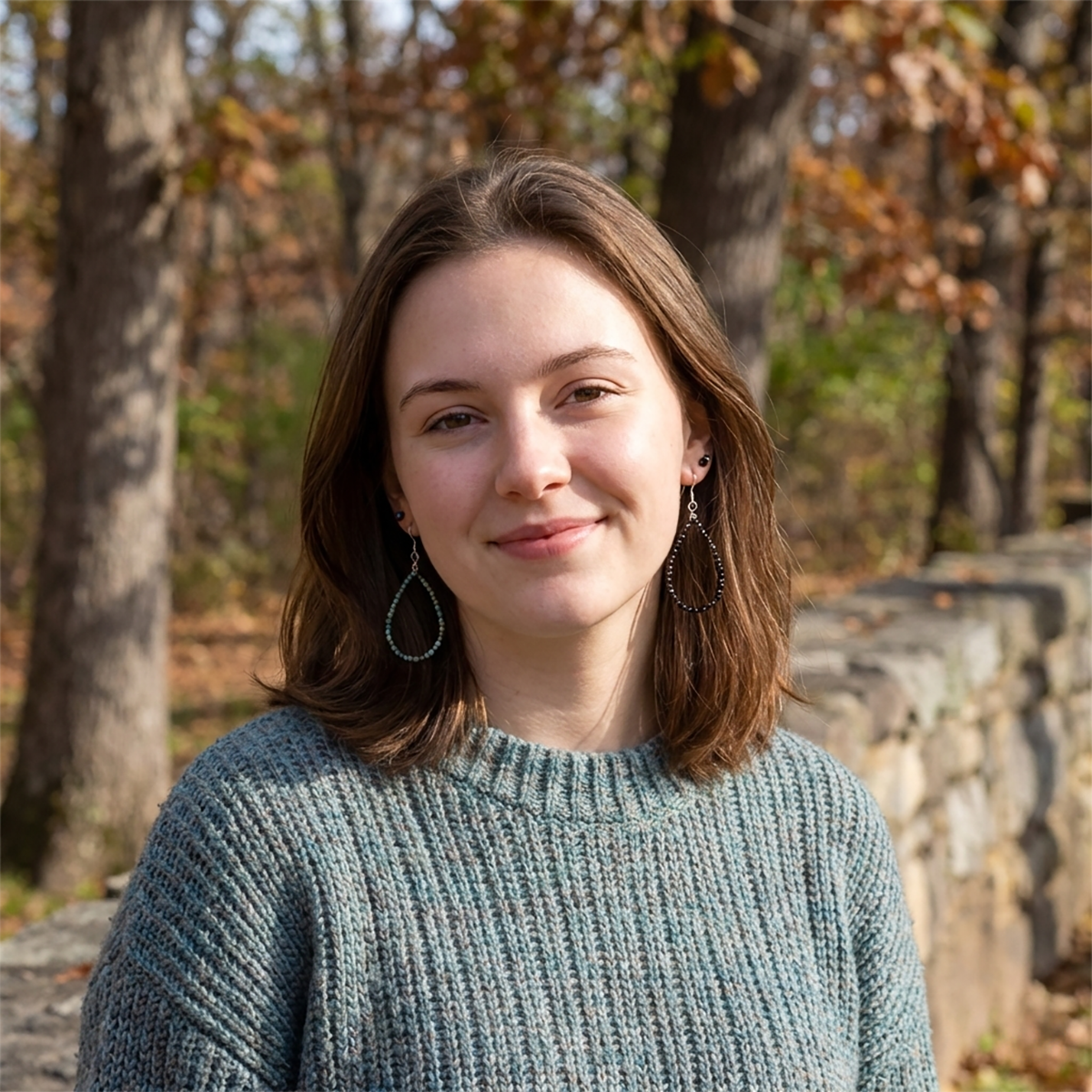 Woman wearing a teal sweater standing in a forest with autumn foliage.