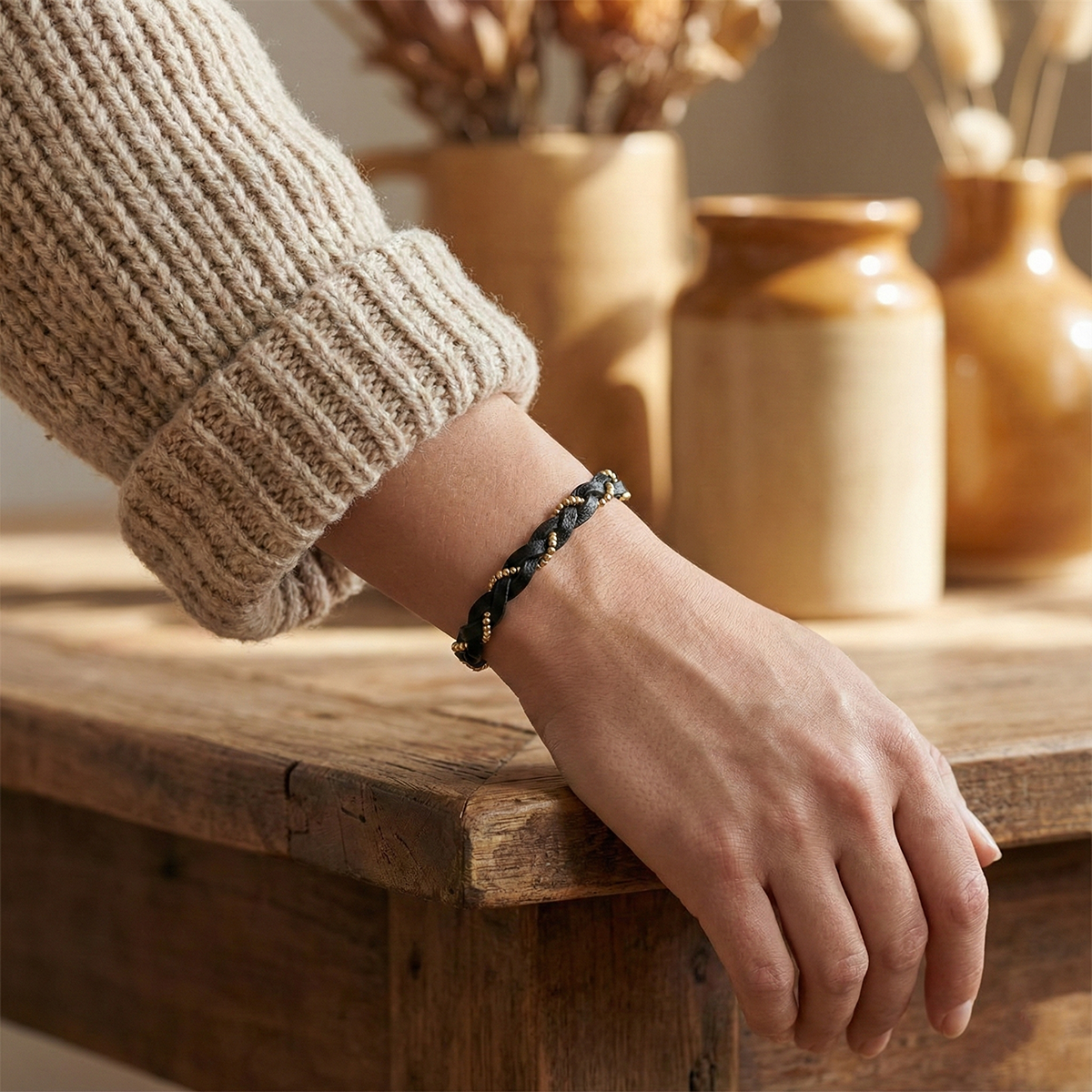 Hand wearing a black braided bracelet on a wooden surface with decorative vases in the background