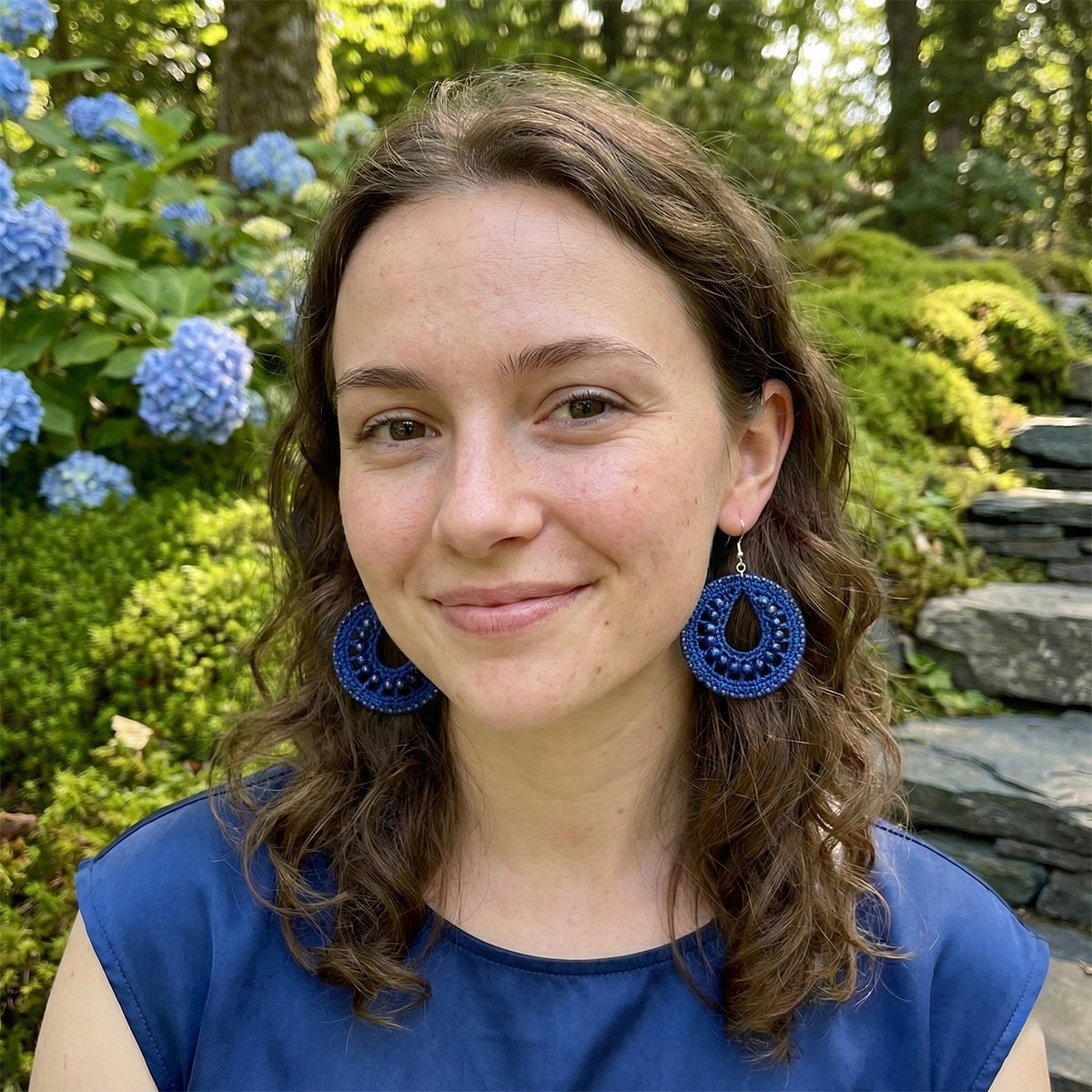 Woman wearing blue earrings in a garden setting with flowers and stones.