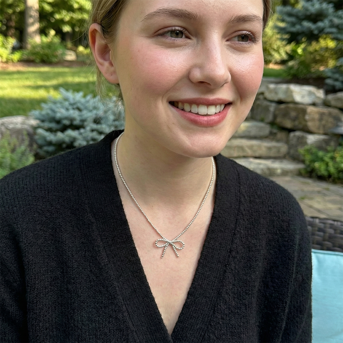 Person wearing a black sweater and a necklace with a bow pendant outdoors.