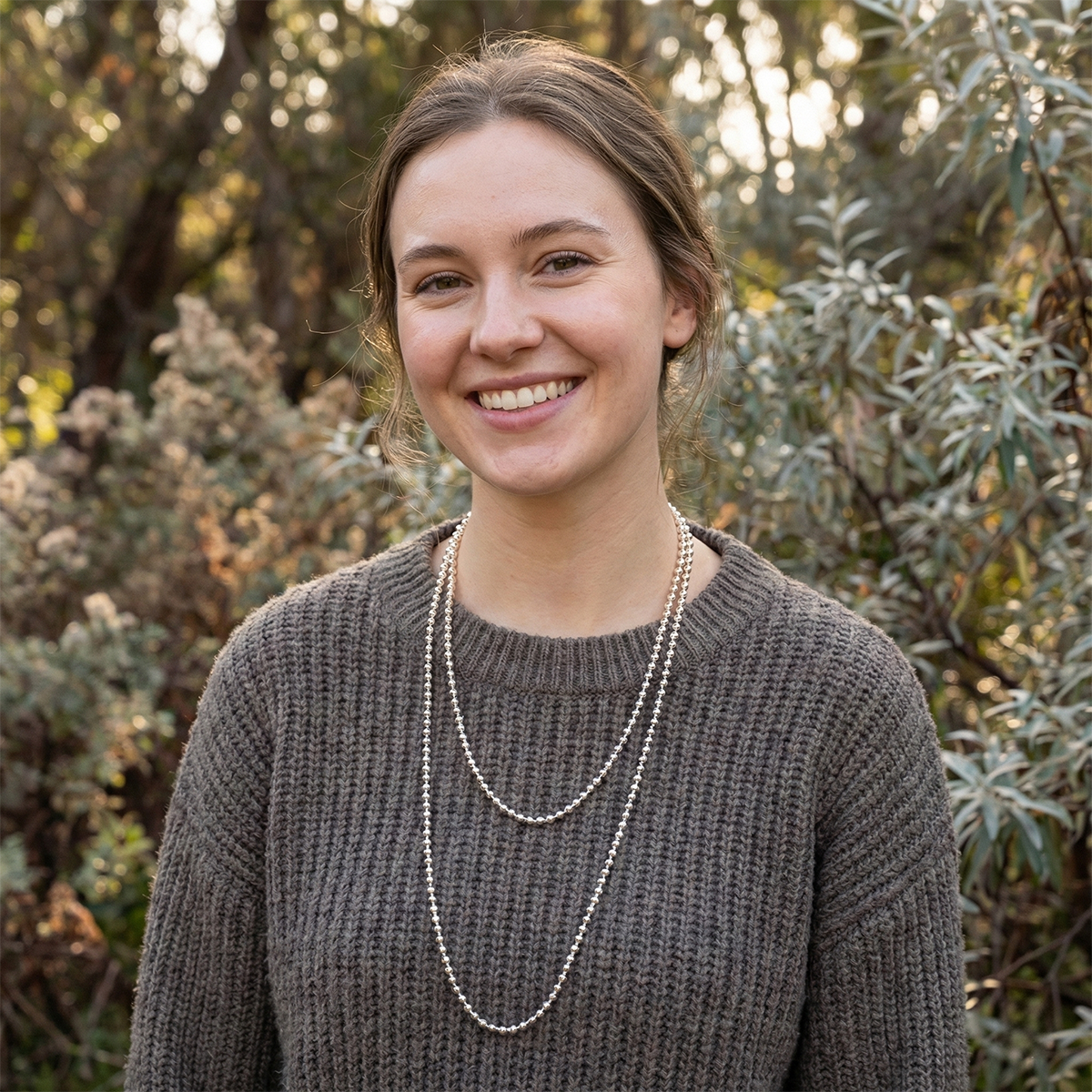 Woman wearing a brown sweater and layered necklace standing in front of greenery